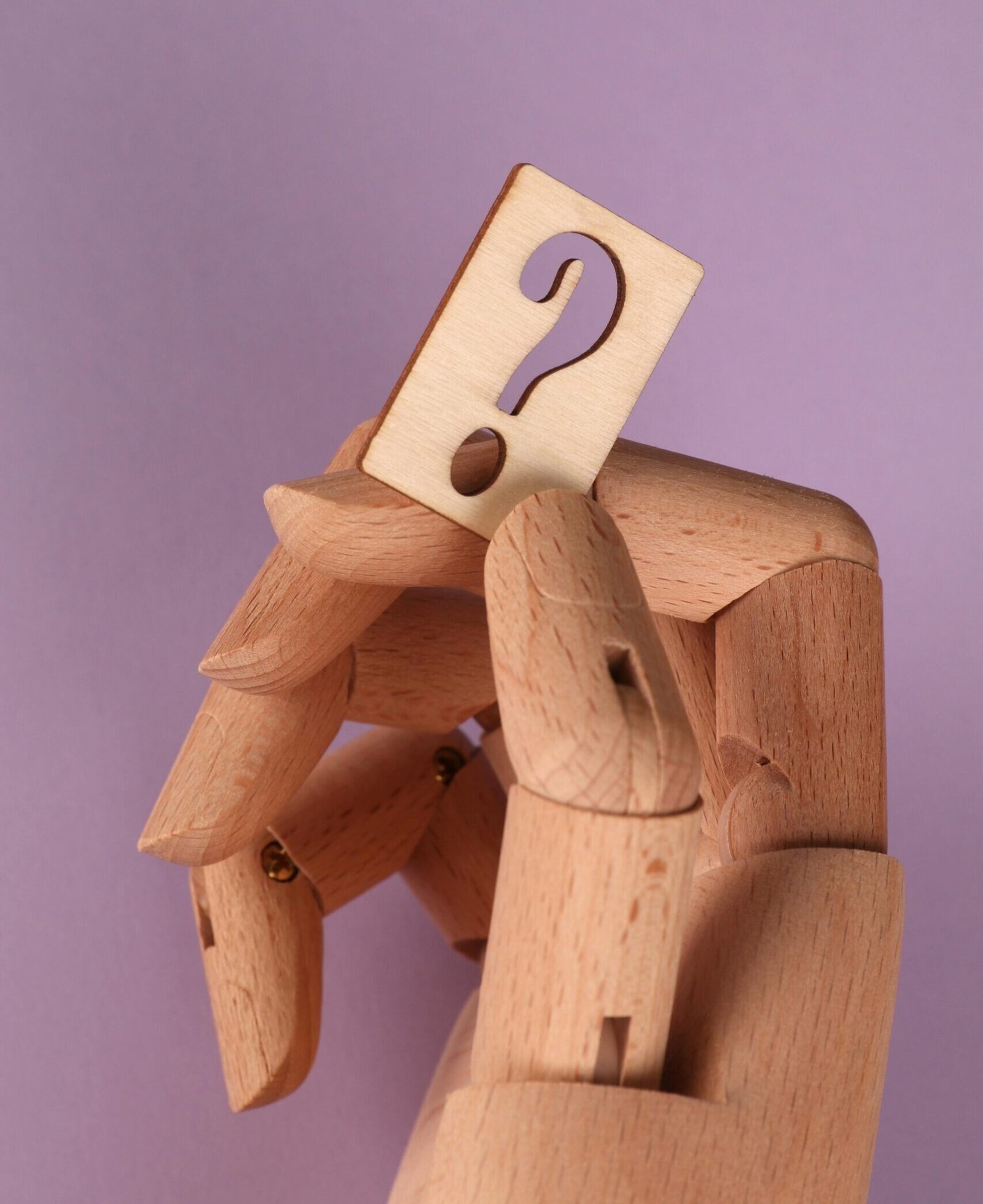 Wooden mannequin hand holding a carved question mark against a purple background.