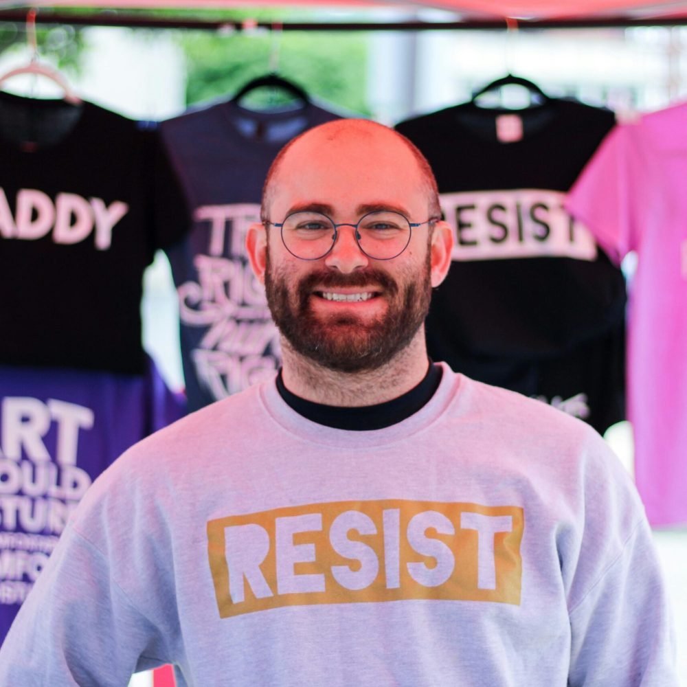 A bearded man wearing a 'Resist' sweatshirt smiles at a market stall showcasing activist-themed shirts.